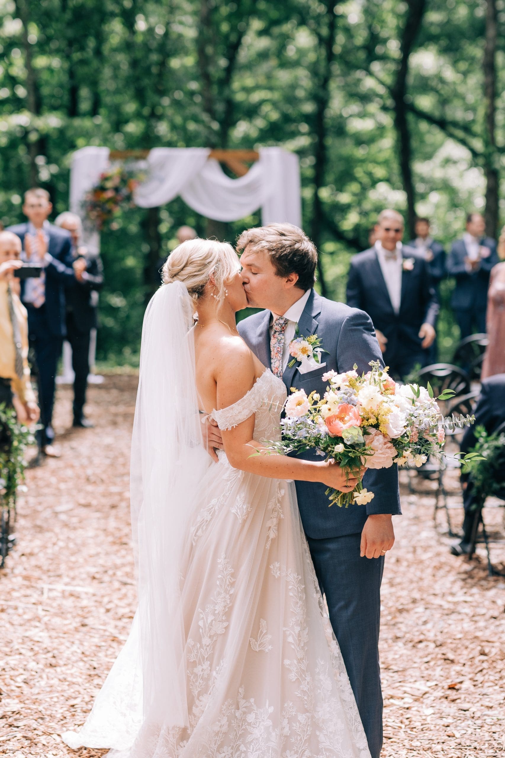 Bride and groom kissing outdoors, surrounded by guests, with a floral arch in the background.