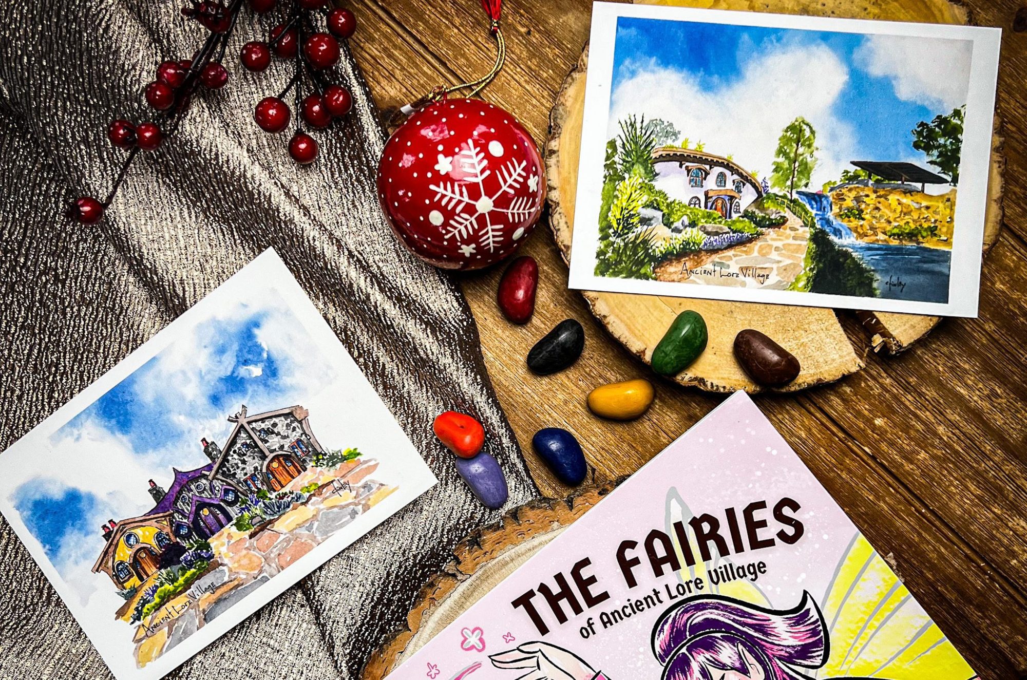 Postcards and a book titled "The Fairies of Ancient Lore Village" are displayed on a wooden surface with some berries and rocks, making an enchanting setup perfect for the gift-giving holidays.