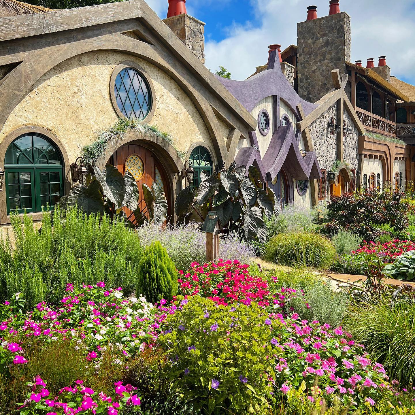 A whimsical, fairy-tale style house with vibrant flowers and plants in the foreground under a bright blue sky.