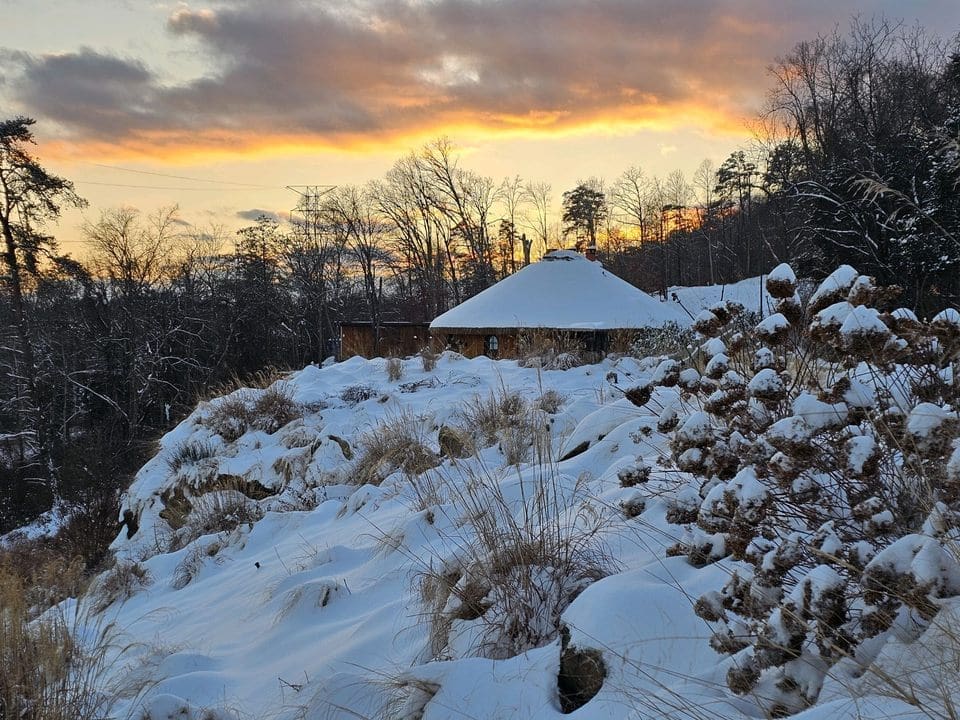 snowy roof A snowy landscape cradles a circular house surrounded by bare trees, where intentional gatherings with friends and family unfold beneath the warm hues of a sunset sky.