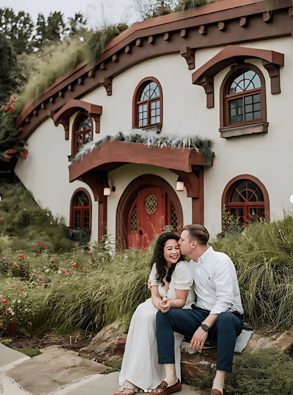 Couple sitting on a bench, embracing in front of a quaint, hobbit-like house surrounded by greenery.