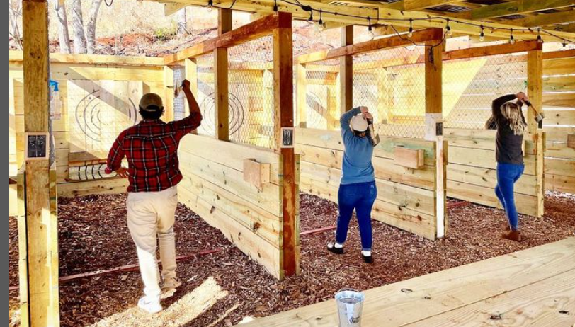 Three people are throwing axes at wooden targets in an outdoor lane with wood chips on the ground, embracing one of the 5 Ways to Start Your New Year With Magic at Ancient Lore Village—a place where every toss adds a touch of enchantment to your fresh beginnings.