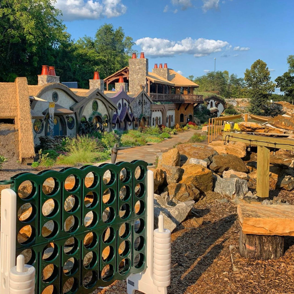 Outdoor scene with a large Connect Four game in the foreground, whimsical hobbit-like houses in the background, and a clear blue sky. Picture laughter echoing as friends gather around, enjoying charcuterie boards and wine while playing yard games under the sun.