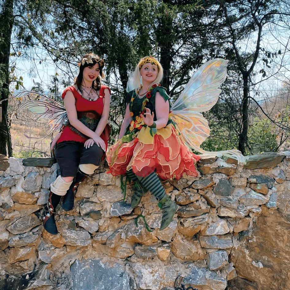 Two people dressed as fairies with wings sit on a stone wall, surrounded by trees and nature, celebrating International Fairy Day.