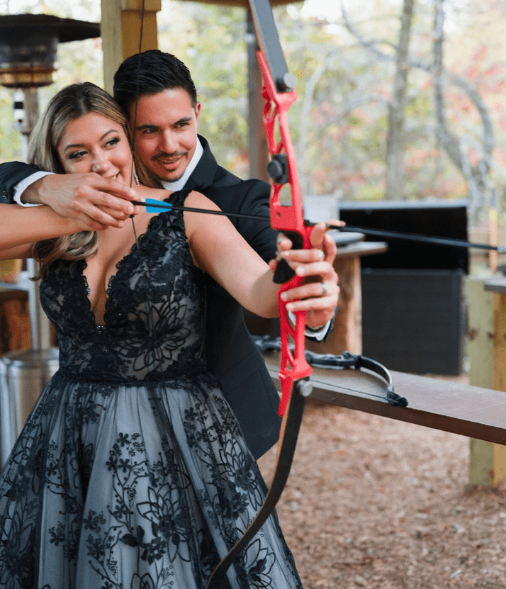 A couple practicing archery together; the woman holds a bow while the man assists her outdoors.